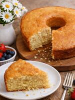 bolo de laranja em uma tábua redonda de madeira, com uma fatia faltando, ao lado um prato brnaco com uma fatia do bolo.