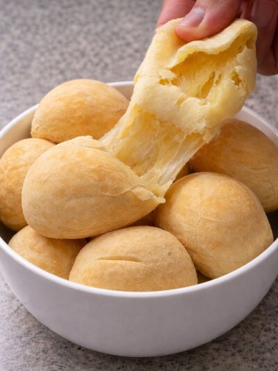 Vários pães de queijo veganos, redondos, um está esticado mostrando a sua elasticidade.