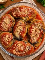 Travessa de vidro com bifes vegetais empanados ao molho de tomate e cobertura crocante, servidos sobre uma mesa de madeira rústica com ervas frescas e toalha de tecido.